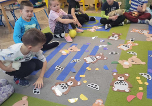 Dzieci siedzą na dywanie. Przed nimi leżą karty wizerunkami misiów trzymających różne kształty, kolory i cyfry.