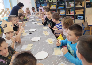 Przedszkolaki siedzą przy stole i jedzą wafle polane słodkim miodem.