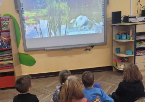 Przedszkolaki siedzą na dywanie i oglądają prezentację PowerPoint na tablicy multimedialnej