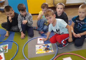Dzieci siedzą na dywanie. Eryk dołożył rekwizyt do obrazka przedstawiającego baśń "Piękna i Bestia". Pięcioro chłopców obserwuje działanie kolegi.