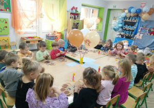 Dzieci siedzą przy złączonych stołach i wykonują pracę plastyczną - misia z torebki papierowej. W tle dekoracja z okazji Dnia Pluszowego Misia.