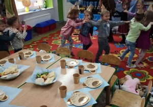 Dzieci podczas zabawy w "pociąg", który prowadzi nauczyciel w stroju misia. Na pierwszym planie stoją złączone stoły, na których leżą niebieskie serwetki, misiowe talerzyki i kubeczki oraz wafelki.