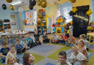 Dzieci siedzą w kole na dywanie podczas zabawy przy piosence "Magiczny Kapelusz". Bartuś trzyma przed sobą kapelusz. W tle dekoracja z okazji Andrzejek, kąciki zainteresowań, a przy suficie wiszą balony.