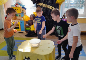 Filip, Kacper, Oskar i Kacper stoją wokół stołu, na którym znajduje się duża miska z wodą i cekinami. Przedszkolaki wrzucają monety do wody.