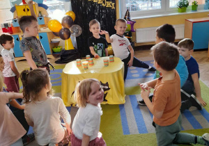Dzieci przykucnęły wokół stolika nakrytego żółtym materiałem, na którym stoją kubki z ukrytymi przedmiotami. Kacper i Oskar wybierają kubki z wróżbą. W tle dekoracja andrzejkowa, kącik domowy, okno.