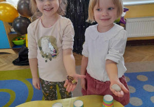 Wiktoria i Łucja stoją obok stolika, na którym stoją kubeczki z wróżbami. Dziewczynki trzymają w dłoniach odkryte przez siebie przedmioty - cukierek i kamień.