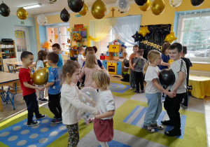 Dzieci tańczą na dywanie z balonami trzymając je między brzuchami. W tle dekoracja andrzejkowa, kącik domowy, przyrodniczy, a przy suficie wiszą balony w kolorze czarnym, złotym i przezroczyste z konfetti.