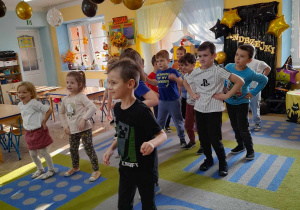 Dzieci wesoło tańczą na dywanie przy muzyce. Nad nimi wiszą balony w kolorze czarnym, złotym i przezroczyste z konfetti. W tle dekoracja andrzejowa, okna, drzwi, stoły.