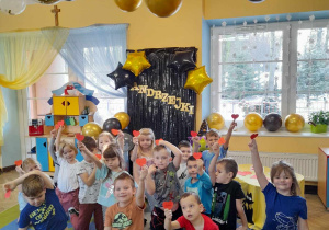 Dzieci siedzą na dywanie. Każde trzyma serduszko w ręku. W tle kącik domowy, dekoracja andrzejkowa, stolik na wróżby, a przy suficie wiszą balony.