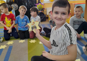 Franek prezentuje obrazek na wylosowanej gwiazdce - Mikołaja. W tle kilkoro dzieci siedzi na dywanie, a za nimi dekoracja andrzejkowa.