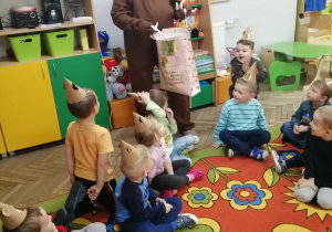 Dzieci siedzą na dywanie w papierowych czapeczkach misia, przed nimi stoi nauczyciel w stroju niedźwiedzia i trzyma w torbie upominek dla dzieci.