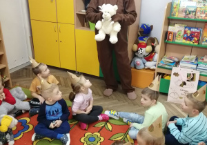 Dzieci siedzą na dywanie w papierowych czapeczkach misia, przed nimi stoi nauczyciel w stroju niedźwiedzia i trzyma pluszowego misia.