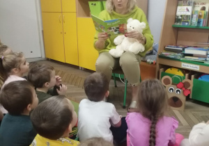 Nauczyciel siedzi trzymając misia i czyta książkę. Przed nim na dywanie siedzą dzieci, w tle kącik książki.