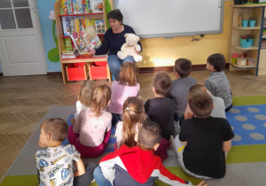 Dzieci siedzą na dywanie, a przed nimi pani Ania czyta dzieciom książkę trzymając Misia Lubisia na kolanach.