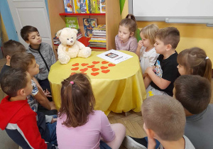 Dzieci siedzą na dywanie wokół stolika nakrytego żółtym materiałem. Na stoliku znajduje się Miś Lubiś, Dzienniczek Małego Misia, serduszka z imionami dzieci. W tle Kącik Książki, tablica multimedialna.