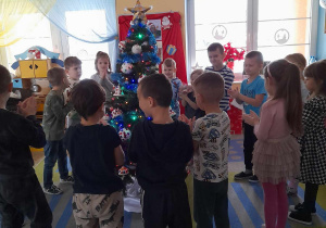 Dzieci stoją w kole na dywanie, a środku ubrana, mieniąca się światełkami choinka. Przedszkolaki śpiewają piosenkę. Nad ich głowami wiszą balony w kolorze złotym, czerwonym i czarnym.