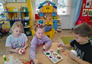 Anastazja, Gabrysia i Julek siedzą obok siebie przy stole. Dzieci ozdabiają kolorowymi elementami styropianowe bombki.