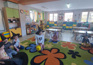Przedszkolaki siedzą w kole na dywanie .W środku kołaJulia rzuca kostką do gry..