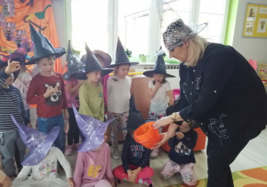 Nauczyciel w czarnym kapeluszu częstuje dzieci z pomaranczowego wiaderka cukierkami.