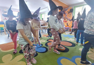 Dzieci w czarnych kapeluszach czarownicy stoją na dywanie w rozsypce i trzymają kolorowe drążki podczas zabawy „Czarownice na miotle”.