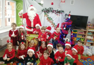 Dzieci z grupy "Motylki" na grupowym zdjęciu z Mikołajem i prezentami na tle dekoracji.