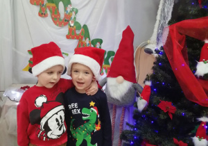Dwóch Mikołajów w czapkach mikołajkowych pozują do zdjęcia na tle dekoracji.