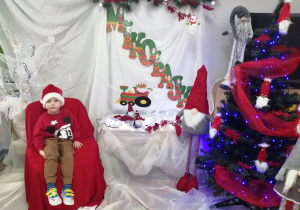 Mikołaj w mikołajkowej czapce siedzi na krześle wyłożonym czerwonym materiałem, W tle mikołajkowa dekoracja.