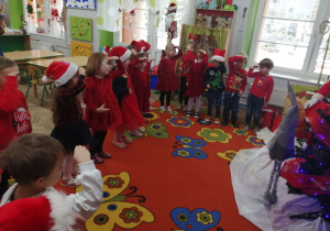 Dzieci w mikołajowych czapkach stoją w półkolu i prezentują piosenkę.