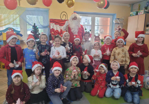 Pamiątkowe zdjęcie Świętego Mikołaja i przedszkolaków z grupy Słoneczek. Dzieci trzymają w dłoniach świąteczne skrzaty – podarunki od Świętego Mikołaja