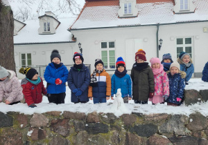 Zdjęcie dzieci z grupy Słoneczek w Parku Wiosny Ludów. Na murku przed przedszkolakami widać ulepionego maleńkiego bałwanka