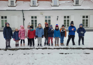 Przedszkolaki z grupy Słoneczek stoją na tle ośnieżonego budynku Szkoły Muzycznej. Część dzieci trzyma w dłoniach ulepione śnieżki