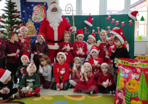 Grupowe zdjęcie dzieci z grupy „Biedronki” z Mikołajem na i prezentami na tle dekoracji.