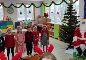 Mikołaj siedzi na tle dekoracji a Adaś, Patryk, Maja, Wiktoria, Mikołaj, Julka i Natalka śpiewają piosenkę. W tle świąteczna dekoracja sali.