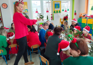 Pani Ania prezentuje gałązki do stroika, przy stolikach: rodzice i dzieci w świątecznych strojach.