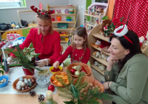 Lena z mamą i Panią Anią ozdabiają świąteczny stroik dodatkowymi elementami.
