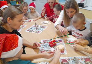Dzieci siedzą z rodzicami przy stolikach i malują lukrem ciastka w kształcie Mikołaja. Na środku stołów leżą serwetki świąteczne.