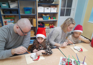 Zosia i Antoś razem z rodzicami malują Mikołaje z ciastek czerwonym i białym lukrem.