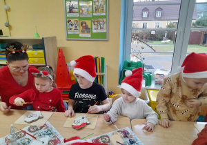 Przy złączonych stolikach siedzi czworo dzieci razem z mamą Gabrysi. Gabrysia i Franek malują pędzelkiem Mikołaja z ciastka.