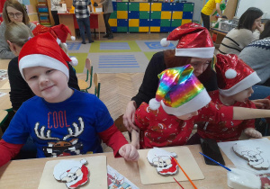 Arek, Samuel i Lucuś siedzą przy stole w czapkach Mikołaja, a przed nimi na deseczkach leżą ciastka w kształcie Mikołaja. Za chłopcami siedzi mama.