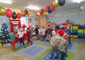 Dzieci ubrane w świąteczne czapki tańczą w parach na dywanie przy piosence o Mikołaju. Nad nimi wiszą przy suficie balony w kolorze złotym, czarnym i czerwonym. Z lewej strony znajduje się ubrana choinka, tablica przykryta czerwonym materiałem z napisem Wesołych Świąt i obrazkiem świątecznym. W tle szafki, okno.
