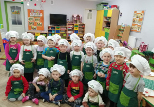 Zdjęcie grupowe dzieci. Przedszkolaki ubrane są w zielone fartuszki i białe czapki kucharskie. W tle szafki z układankami.