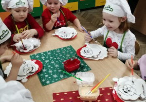 Grupka dzieci przy stolikach maluje kolorowym lukrem ciasteczkowego Mikołaja.