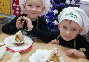 Mikołaj i Marcel prezentują swoje muffinkowe, zimowe domki. W tle ubrana choinka.