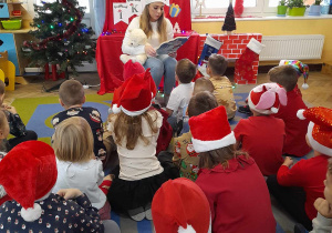 Dzieci siedzą na dywanie, a przed nimi Pani logopeda czyta książkę. Na kolanach trzyma Misia Lubisia. W tle dekoracja z okazji Mikołajek.