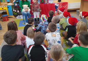 Dzieci siedzą na dywanie, a przed nimi zaproszeni goście. Amelka trzyma książkę, a mama Tymka rozmawia z dziećmi na temat przeczytanego wiersza.