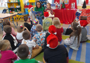 Przedszkolaki siedzą na dywanie. Mama Tymka czyta dzieciom książkę, a obok niej siedzi Amelka, która trzyma na kolanach maskotkę - Misia. W tle dekoracja świąteczna.