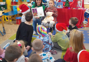 Na dywanie siedzą przedszkolaki, a przed nimi rodzic prezentuje obrazek w książce. Obok mamy siedzi Amelka z Misiem Lubisiem. W tle dekoracja świąteczna.
