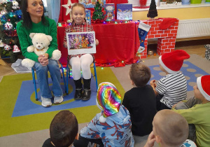 Amelka prezentuje obrazek w książce "Gwiazdkowe wierszyki", a obok siedzi mama Tymka z Misiem Lubisiem na kolanach. Przed zaproszonymi gośćmi siedzą na dywanie dzieci. W tle dekoracja świateczna.