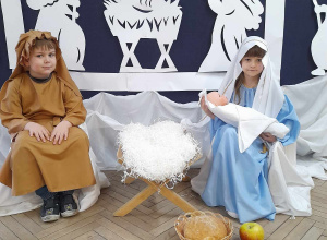 Zosia w roli Maryi i Kacper w roli Józefa siedzą przy żłóbku, a przed nimi leżą dary: bochenek chleba, jabłko, mirra, kadzidło i złoto. w tle fragment dekoracji jasełkowej.