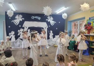 Aniołki i gwiazdki tańczą na środku sceny przy piosence "Przy kominie świerszczyk spał". W tle dekoracja jasełkowa.